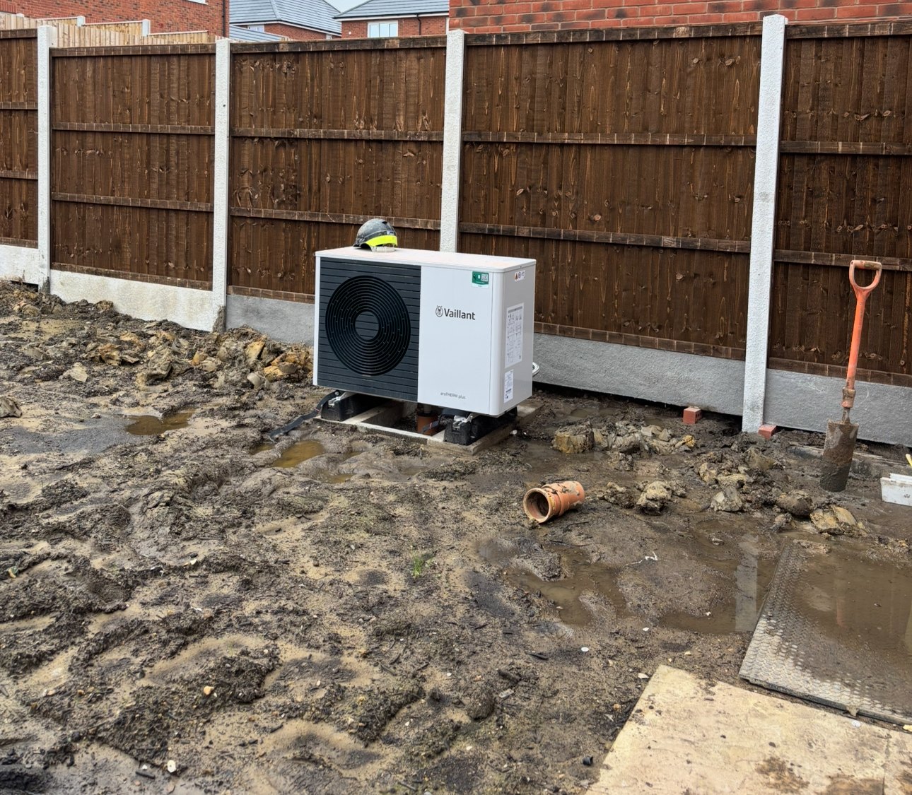 Uncovered heat pump standing in mud with a hard hat left on top